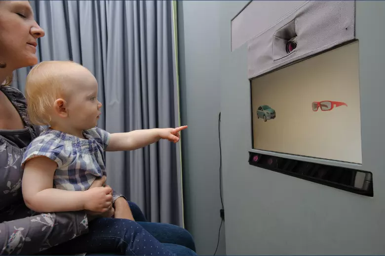Baby sitting on woman's lap, pointing at screen showing sunglasses and a car